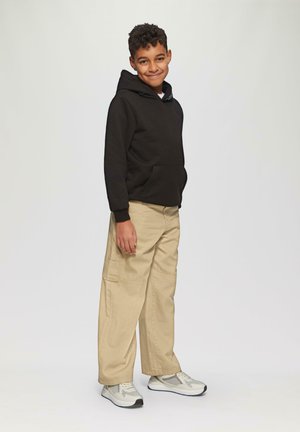 Lächelnder Junge steht, trägt einen schwarzen Hoodie, beigen Cargo-Hosen und weiße Sneaker vor einem einfachen hellen Hintergrund.