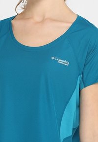 Teal kortärmad Columbia Montrail atletisk skjorta som bärs av en person, visar överkropp och logotyp på vänster bröst.