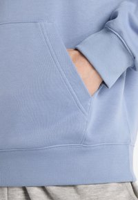 Felpa azzurra chiaro con tasca a marsupio, polsini e orlo a coste. Caratterizzata da una trama di tessuto morbido e dettagli di cucitura visibili.