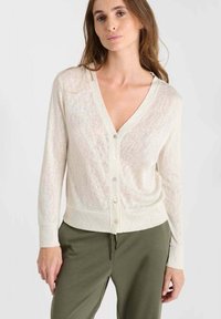 Hellbeige Cardigan mit V-Ausschnitt, langen Ärmeln und Knopfleiste. Strukturiertes Gewebe mit weichem Finish, kombiniert mit olivgrünen Hosen.