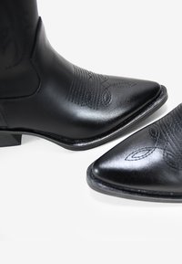 Stivali da cowboy in pelle nera con punta affusolata, caratterizzati da motivi di cucitura decorativi. Il design include un tacco basso e una superficie liscia.