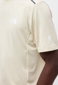 Μπεζ κοντομάνικη μπλούζα που φοράει άτομο με σκουρόχρωμο δέρμα, που έχει λευκό λογότυπο "The North Face" στο στήθος και λογότυπο "Mountain Athletics" στο μανίκι.