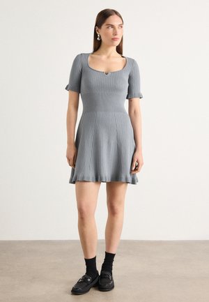 Light grey ribbed dress with a square neckline, short sleeves, flared skirt, and a ruffled hem. Styled with black socks and loafers.