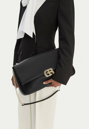 Person in schwarzem Blazer und weißer Bluse hält große schwarze Leder-Clutch mit goldener Kettentasche und goldenem Doppel-G-Logo.
