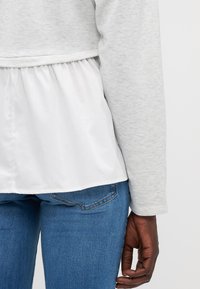 Sudadera gris claro con parte inferior plisada blanca. La textura es suave, con mangas largas y un ajuste relajado, combinada con jeans de mezclilla azul.