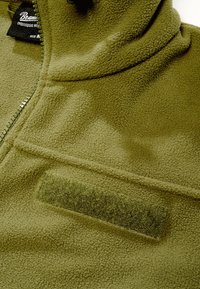 Gros plan d'une veste polaire vert olive avec une fermeture éclair, une patte Velcro et une étiquette de marque noire à l'intérieur du col.