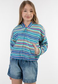 Giacca bomber a motivi multicolor con strisce blu, rosa e verde, dotata di zip frontale e tasche laterali, abbinata a pantaloni shorts in denim.
