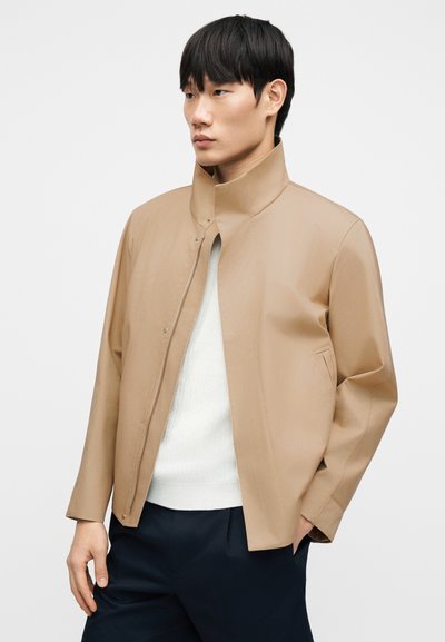 Veste beige légère avec un col montant, fermeture à pression et poches latérales. Portée par-dessus un pull blanc et un pantalon foncé, présentant une texture lisse.