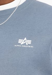 Šviesiai mėlynas medvilninis džemperis. Priekyje yra baltas logotipas su trimis horizontaliais dryžiais. Guma kaklo ir rankovių dalyje.