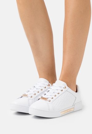 Sneaker bianchi basse con pannelli laterali testurizzati e dettagli in metallo dorato indossati ai piedi di una persona su uno sfondo bianco.