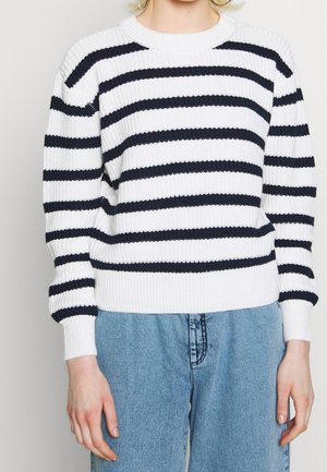 Personne portant un pull blanc en tricot à rayures horizontales noires et un jean bleu en denim, debout avec les bras relâchés le long du corps.