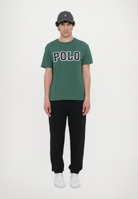 Groene katoenen T-shirt met "POLO" in witte en marineblauwe letters, gecombineerd met zwarte joggingbroeken en witte sneakers. De pet is donkergrijs met logo.