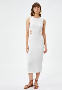 Witte, geribbelde midi-jurk met uitsnijdingen in de taille. Ontworpen met een ronde halslijn en een aansluitende silhouette. Gepaarde met bruine sandaaltjes met bandjes.