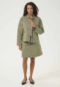 Chaki žalia striukė su mygtukų priekine dalimi ir kišenėmis, derinama su atitinkama sijonu. Medžiaga atrodo kaip medvilnė; švelnus paviršius. Juodi smailėjančio profilio bateliai.