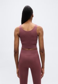 Γυναίκα με θέα από πίσ ω που φοράει μωβ ριμπ αθλητικό σουτιέν και αντίστοιχο κολάν με ψηλή μέση σε μονόχρωμο φόντο.