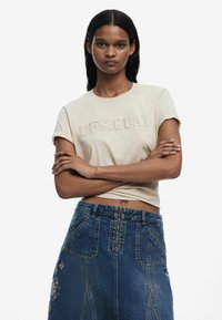 T-shirt in cotone beige con la scritta "DESIGUAL" in rilievo; maniche corte. Indossato con una gonna in denim blu a vita alta con dettagli floreali ricamati.