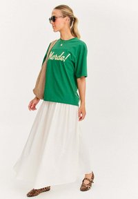 Zelené bavlnené tričko s nápisom "Merde!" v svetložltej farbe, skombinované s voľnou bielou sukňou a balerínami s leopardím vzorom. Slnečné okuliare a sieťovaná taška sú zahrnuté.