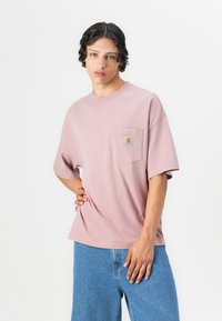 Svetlo roza majica s kratkimi rokavi in žepom na prsih, ki vsebuje logo. Ohlapen kroj, mehak material, kombinirana z modrimi jeans kratkimi hlačami.