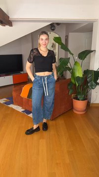 Ung kvinne står i stuen iført svart blonder crop top, blå jeans med snorer og svarte loafers, med en stor potteplante i nærheten.