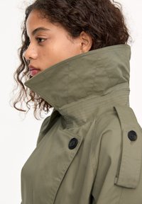Olijfgroen trenchcoat met een hoge kraag, zwarte knopen, gewatteerde accenten en schouderepauletten, die een gladde stofstructuur toont.