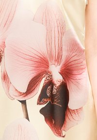 Gros plan sur un tissu avec un grand motif de fleur d’orchidée rose et rouge foncé sur un fond beige clair, montrant les nervures et textures détaillées des pétales.