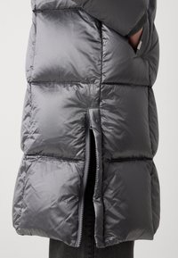 Sivá prešívaná bunda s lesklým povrchom, ktorá má bočné zapínanie na zips a rebrové manžety. Materiál sa javí ako ľahký a izolovaný.