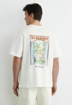 Homme portant un t-shirt blanc avec un motif coloré de fenêtre ouverte comportant des palmiers et le texte "LA HABANA" dans le dos.