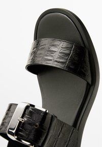 Sort sandal med en tekstureret falsk krokodilleskindstropp, metalspænde og polstret sål med glat overflade.