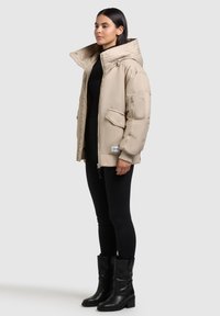 Beige puffjacka med hög krage, dragkedja, sidofickor och vadderat foder. Stylo med svarta leggings och ankelskor.