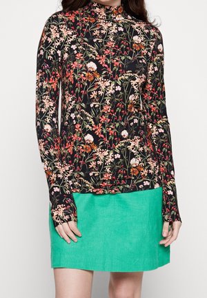 Femme portant un top noir à manches longues avec un imprimé floral coloré et une mini jupe en velours côtelé vert vif, posant devant un fond blanc.