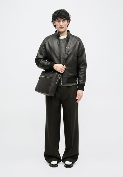 Chaqueta bomber de cuero negro, suéter gris, pantalones oscuros holgados, llevando un bolso mensajero de cuero oscuro con cierre de cremallera y correa ajustable.