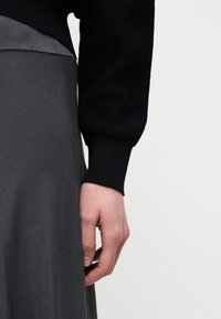 Main détendue le long du corps portant un haut à manches longues noir avec un poignet côtelé et une jupe texturée gris foncé sur un fond clair uni.