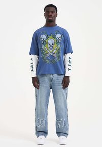 Blaues Grafik-T-Shirt mit Schädel- und Schlangenmotiv, kombiniert mit einem langen weißen Ärmel. Ausgebleichte Jeans mit leichter Waschung und bestickter "1971"-Aufschrift.