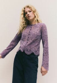 Lange Spitzenoberteil mit langen Ärmeln in Helllila, mit gewelltem Saum, Knopfleiste und zarten Blumenmustern. Kombiniert mit dunklen Jeans.