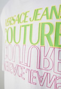 Bílý bavlněný tričko se silným, překrývajícím se textem v zelené a růžové barvě, který zní "VERSACE JEANS COUTURE" v moderním stylu písma.