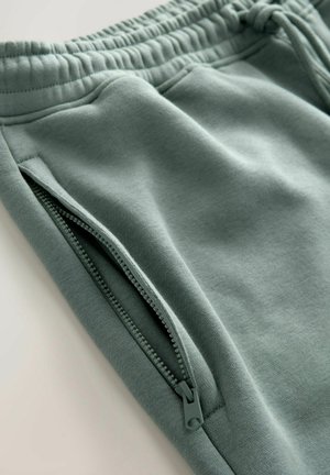 Pantalones de chándal de color verde claro fabricados con una tela suave, con una cinturilla elástica y un detalle de bolsillo lateral con cremallera. Textura suave, diseño informal.
