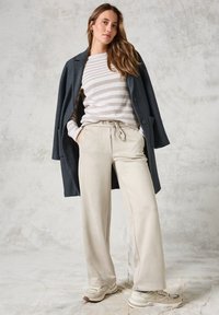 Cappotto lungo grigio scuro su un maglione a righe beige e bianche, abbinato a pantaloni beige con coulisse e sneakers chiare. Texture lisce ovunque.