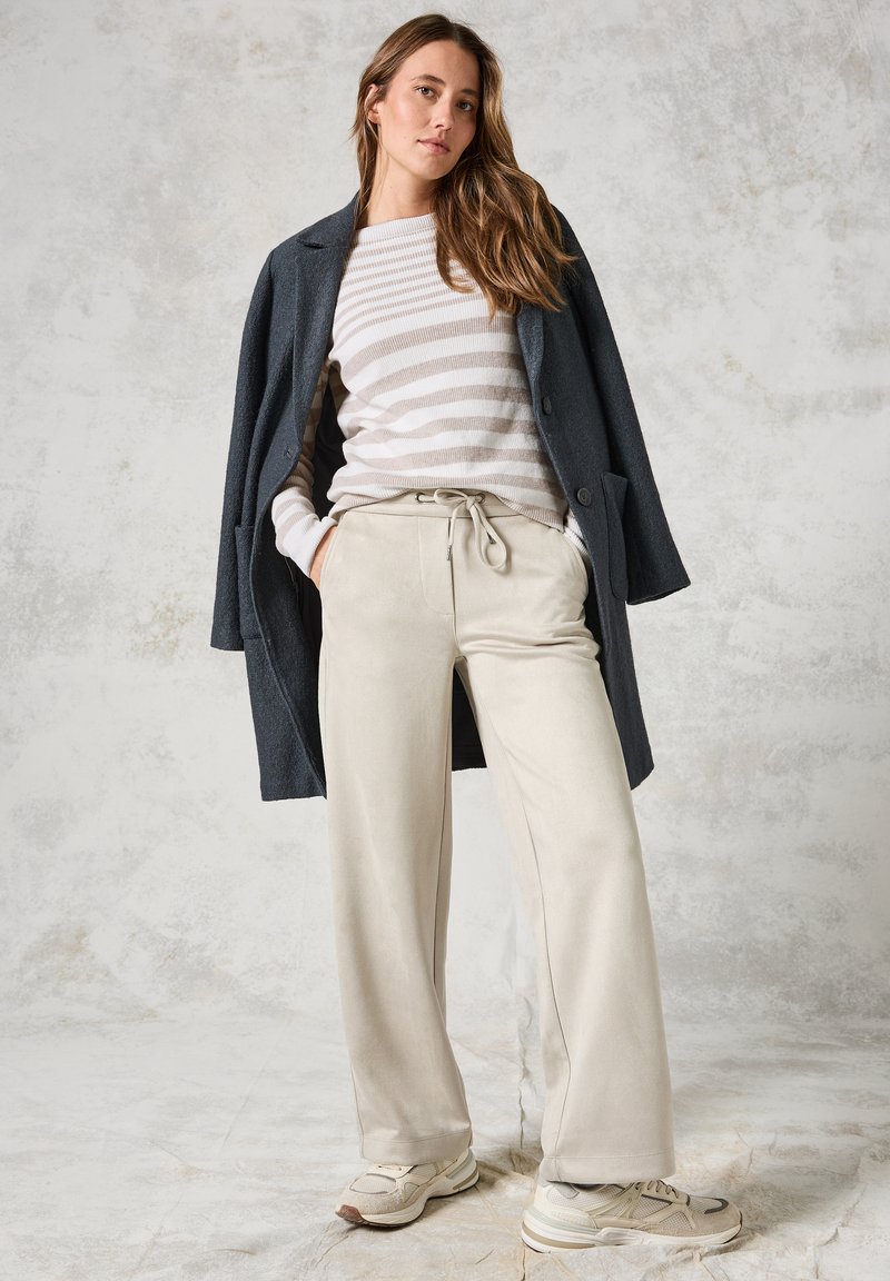 Cappotto lungo grigio scuro su un maglione a righe beige e bianche, abbinato a pantaloni beige con coulisse e sneakers chiare. Texture lisce ovunque.