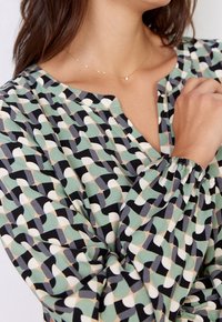 Blusa a maniche lunghe con un motivo geometrico multicolore che presenta tonalità di verde, nero e crema. Tessuto morbido con polsini arricciati e una placca sul collo.