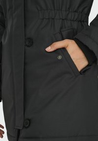 Parka isolante noire avec fermeture à boutons, taille élastique et une poche latérale. Présente une texture lisse et un design minimaliste.