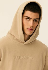 Beżowa polarowa bluza z kapturem, z kieszenią kangurzą i wytłoczonym logo "Diverse Athletics" z przodu. Miękka w dotyku z dużym kapturem.