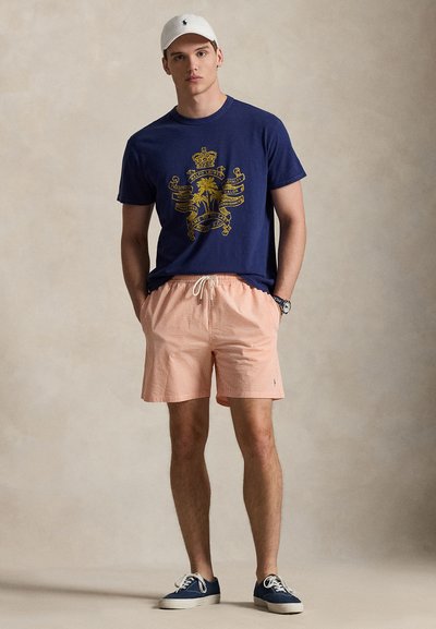 Μπλε μπλουζάκι με χρυσό γραφικό, ανοιχτό πορτοκαλί ριγέ σορτς και λευκό καπέλο. Μπλε αθλητικά παπούτσια ολοκληρώνουν την εμφάνιση. Casual στυλ με άνετη εφαρμογή.