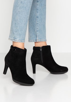 Schwarze Wildleder-Stiefeletten mit seitlichen Reißverschlüssen und Blockabsätzen, getragen zu hellblauen, gekürzten Jeans auf weißem Boden.