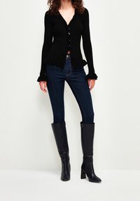 Czarny prążkowany kardigan z dekoltem w serek, zapinany na guziki, z falbaną na dole, zestawiony z ciemnoniebieskimi dżinsami i czarnymi, sięgającymi kolan, obcasowanymi kozakami.
