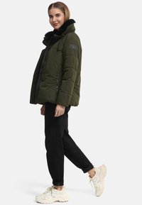 Puffer jacket in olive green with a faux fur collar, zippered pockets, and a slightly loose fit, paired with black pants and light sneakers.