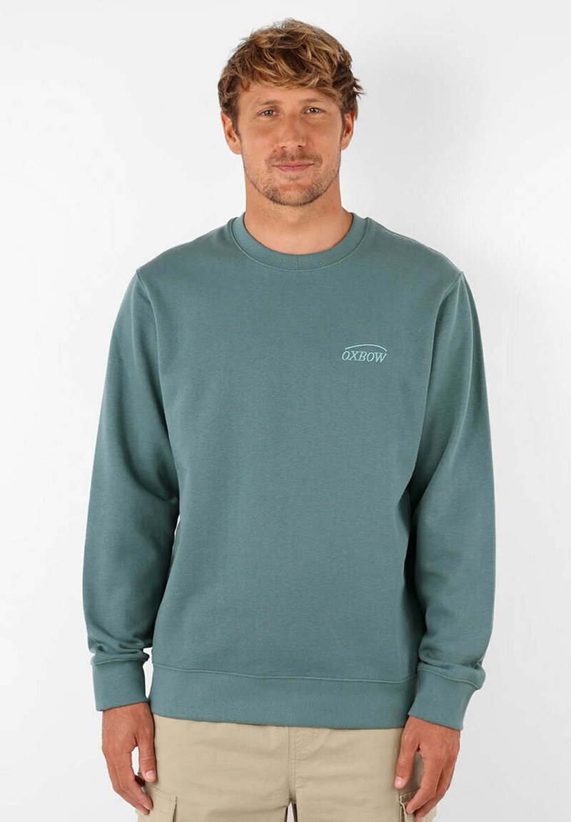 Sweatshirt bleu-vert clair en mélange de coton, avec un col rond, des poignets et un ourlet côtelés, et un logo discret sur la poitrine gauche. Texture lisse.
