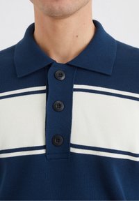 Polo shirt navy con colletto, tre bottoni neri e strisce orizzontali bianche sul petto. Tessuto morbido e testurizzato.