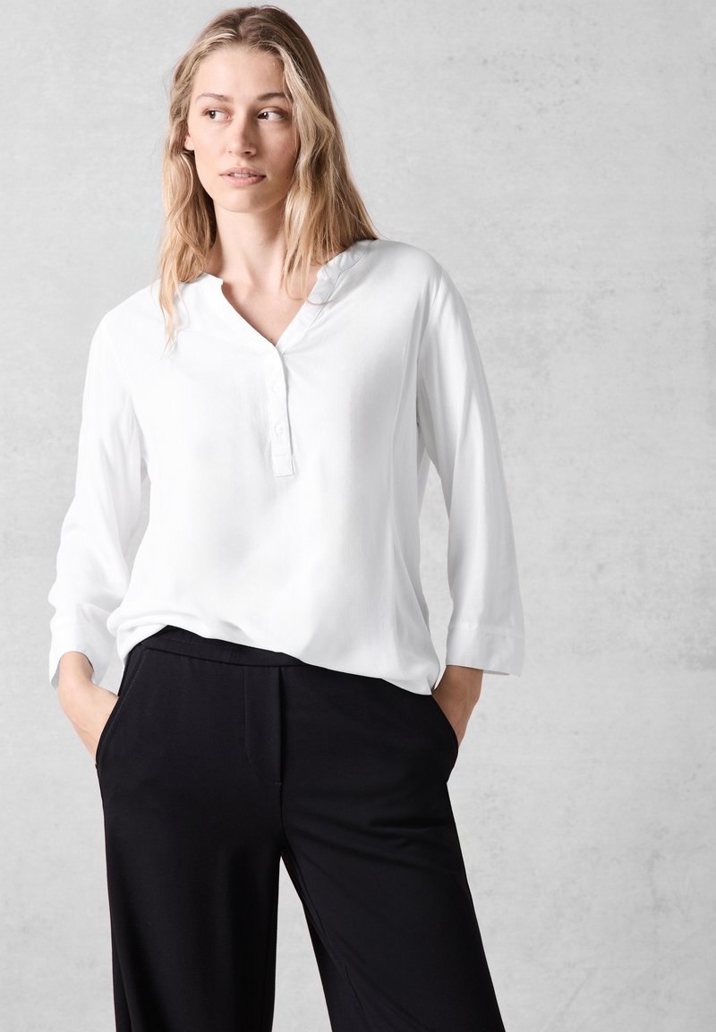 Blusa blanca de manga larga con botones, escote en V y textura suave, combinada con pantalones negros con bolsillos laterales. Caída sutil y ajuste relajado.