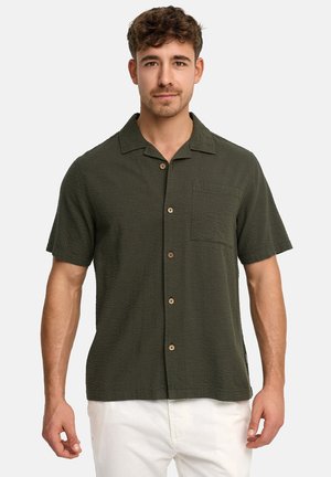Mężczyzna w ciemnozielonej, teksturowanej koszuli na krótki rękaw z kieszenią na piersi oraz w białych spodniach, stojący na jednolitym tle.