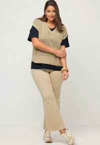 Beige gestrickte Weste mit V-Ausschnitt über einem dunkelblauen T-Shirt, kombiniert mit hellbeigen Schlaghosen und beigen Turnschuhen. Texturiertes und lässiges Design.